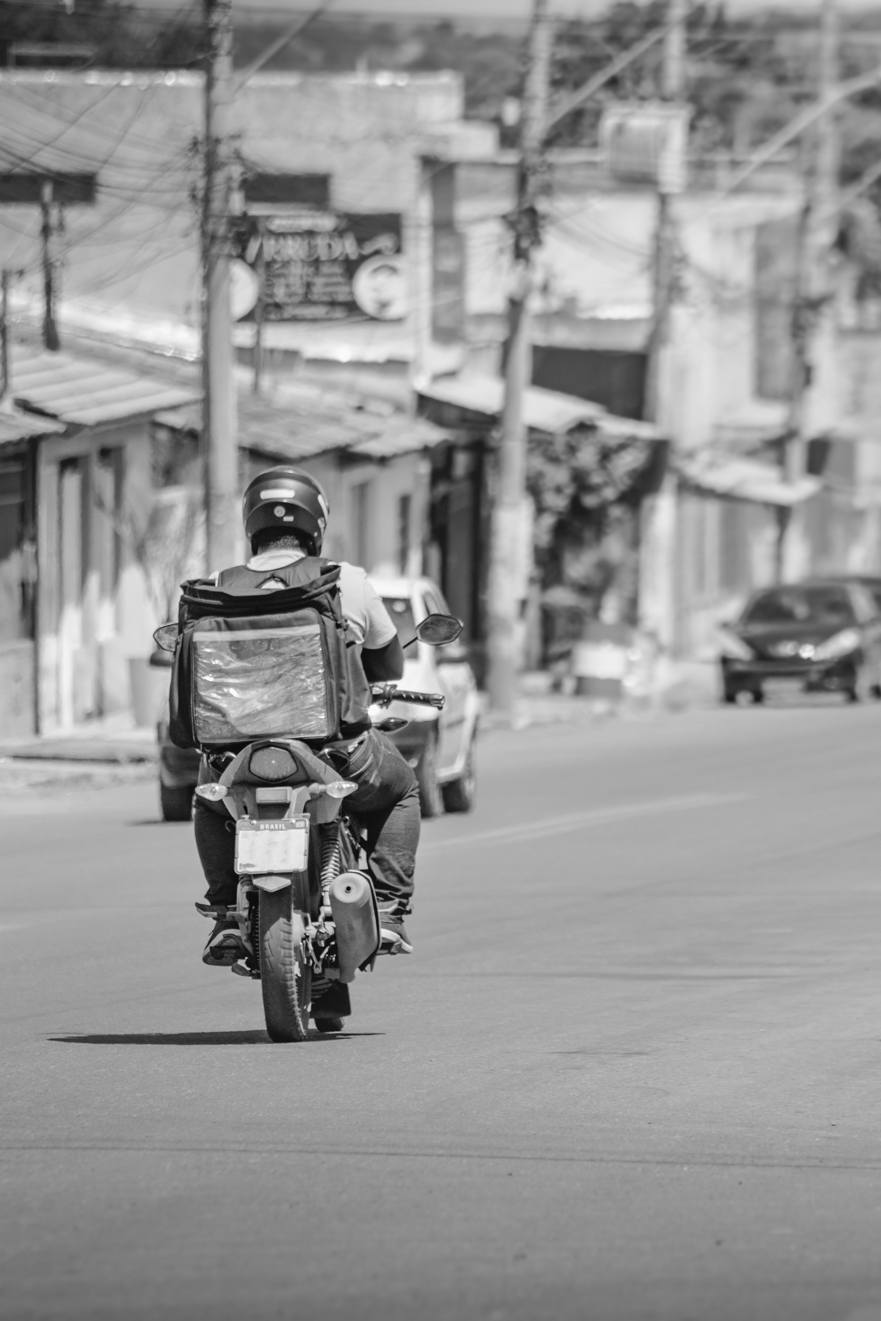 Motociclista entregador de aplicativo visto de costas, pilotando pela rua com mochila térmica de entrega, placa BRASIL na moto